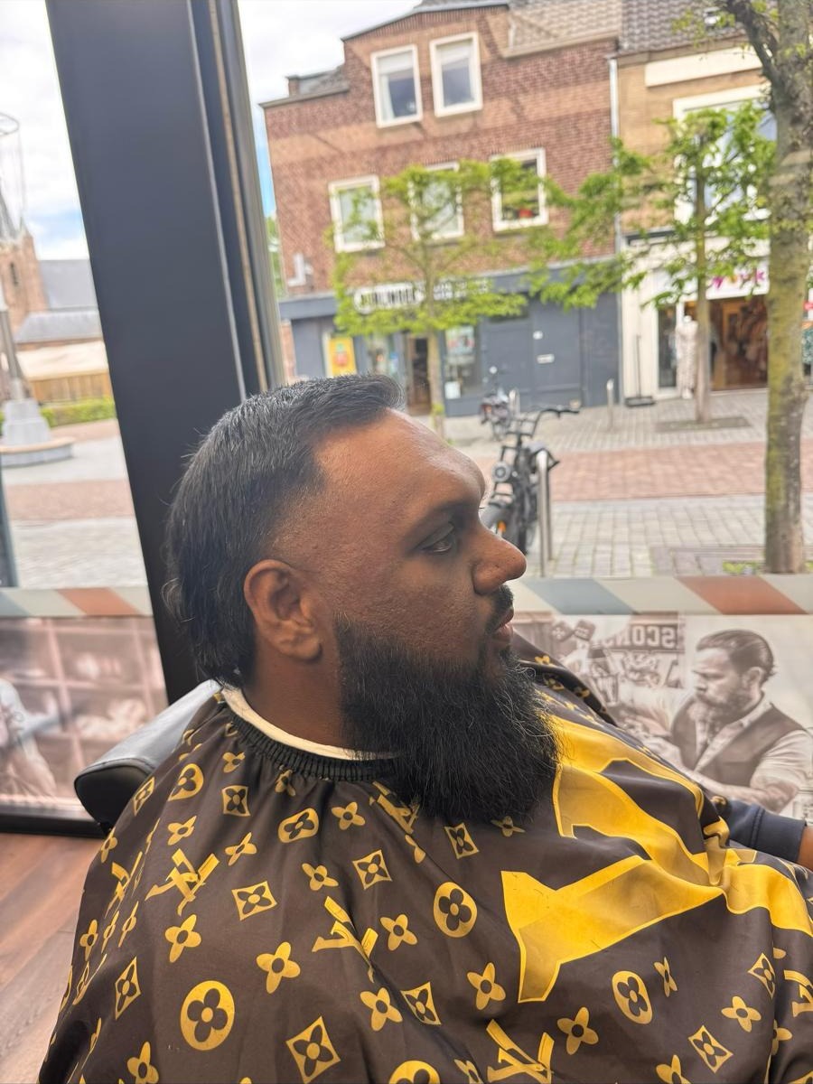 Klant met een strakke fade en een lange zwarte baard in de stoel bij het raam, met de Hoofdstraat erachter