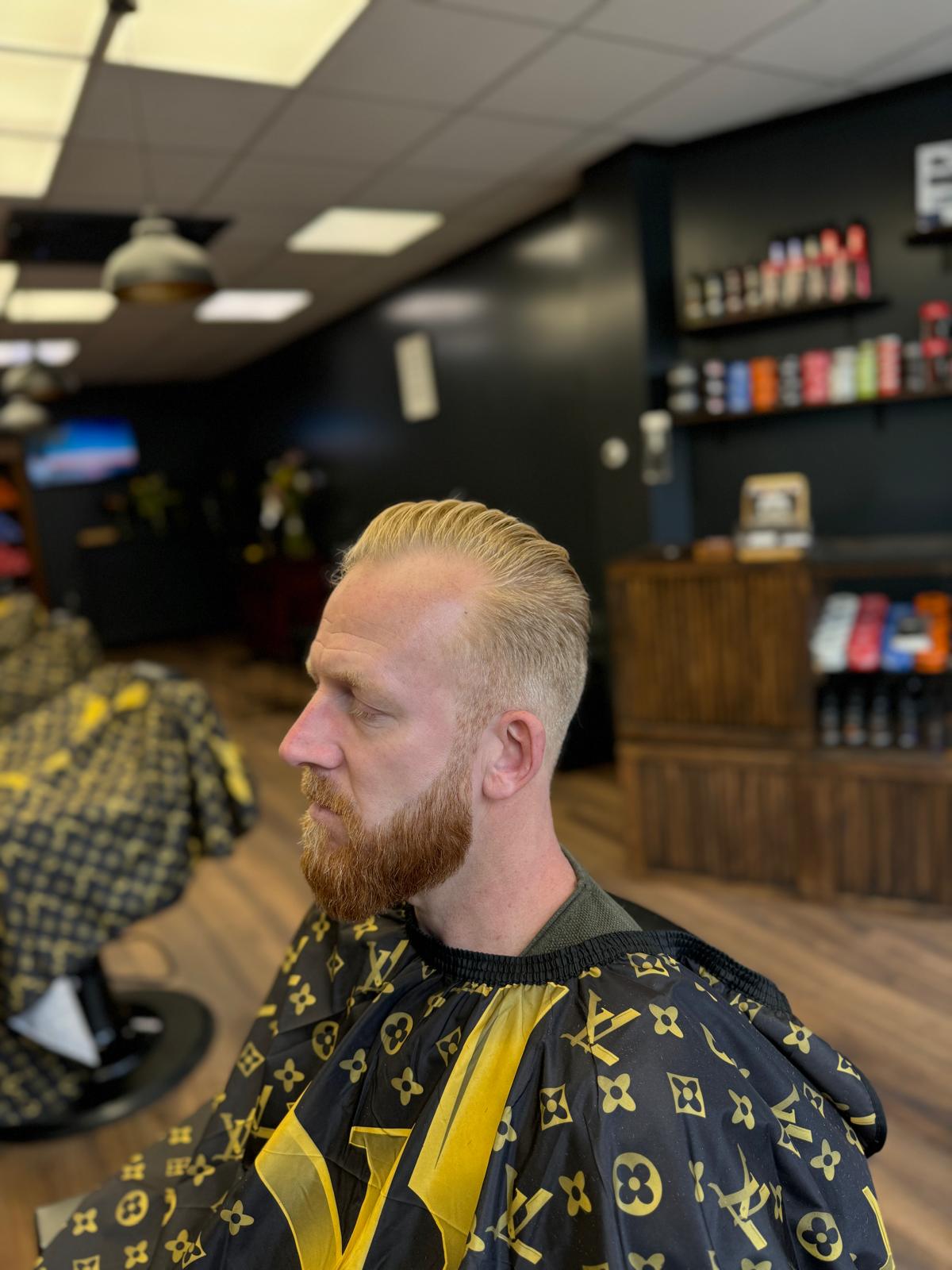 Klant met een blonde fade en een volle baard in de kappersstoel, met de houten wand en de productplanken van de zaak erachter